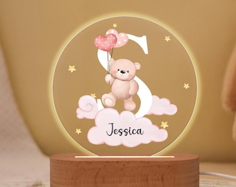 May include: A round, illuminated night light with a wooden base. The design features a teddy bear holding heart-shaped balloons, the letter "S", clouds, and the name "Jessica". The light emits a warm glow.