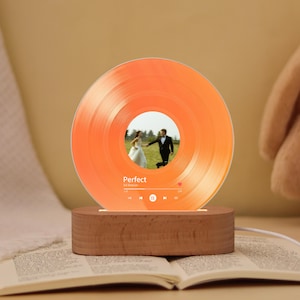 May include: An orange vinyl record-shaped lamp with a photo of a couple in the center. The lamp is on a wooden base and has the word "Perfect" and music controls printed on it. The lamp is lit up and sitting on top of an open book.