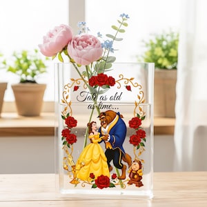 Könnte beinhalten: Transparente Acrylvase mit floralem Design und dem Text "Tale as old as time...". Die Vase zeigt eine Illustration von Belle und dem Biest, umgeben von roten Rosen und goldenen Akzenten. Rosa Pfingstrosen und blaue Blumen sind im Inneren.
