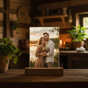 Op de afbeelding: Een rechthoekige fotolijst met een warm, gloeiend licht, met een knuffelend stel, geplaatst op een houten voet. De afbeelding toont een man en een vrouw die een boeket bloemen vasthouden, met een zonsondergang op de achtergrond. De lijst staat op een houten tafel.