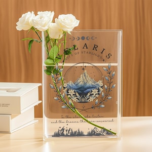 Puede incluir: Un jarr&oacute;n rectangular transparente con un dise&ntilde;o floral y el texto "FLARIS CITY OF STARLIGHT". El jarr&oacute;n contiene rosas blancas y tallos verdes. El dise&ntilde;o incluye una escena de monta&ntilde;a y las palabras "To the stars who listen and the dreams that are answered."