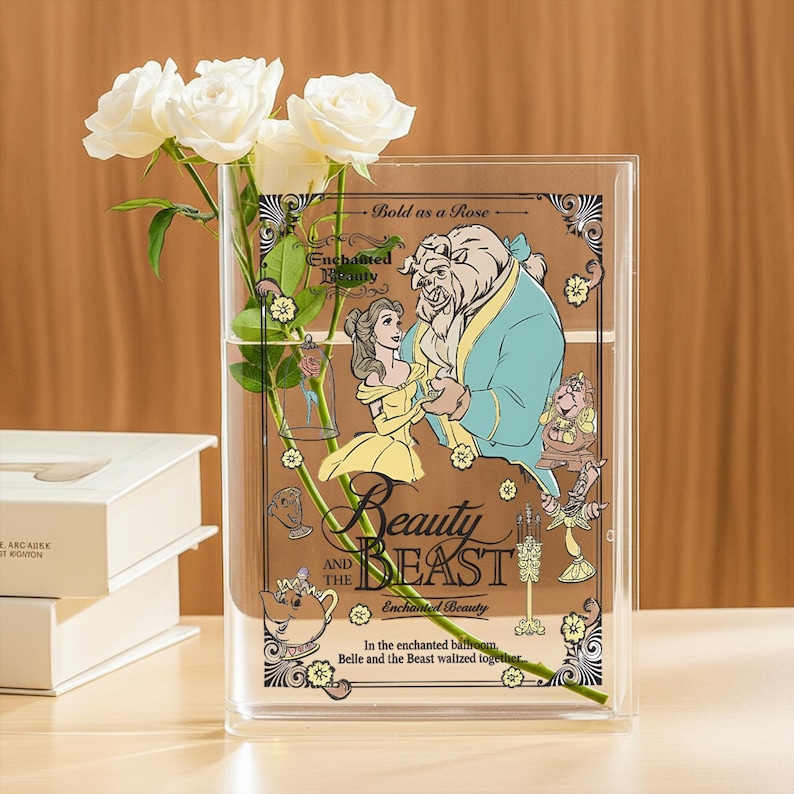 May include: Clear rectangular vase with a Beauty and the Beast design. The vase features Belle and the Beast, with the words "Beauty and the Beast" and "Bold as a Rose." White roses are inside the vase.