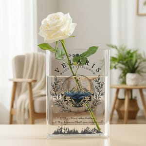Puede incluir: Un jarr&oacute;n rectangular transparente con el texto "VELARIS CITY OF STARLIGHT" y una ilustraci&oacute;n de una escena de monta&ntilde;a. Una rosa blanca con hojas verdes se coloca dentro del jarr&oacute;n. El jarr&oacute;n est&aacute; lleno de agua.