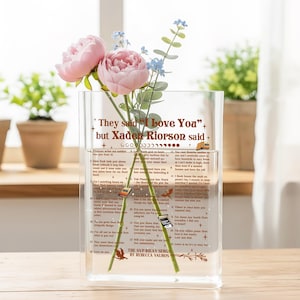 Puede incluir: Un jarrón rectangular de vidrio transparente con texto y un arreglo floral. El jarrón contiene peonías rosas y flores azules. El texto en el jarrón dice "They said 'I Love You' but Xaden Riorson said..." con texto adicional debajo.