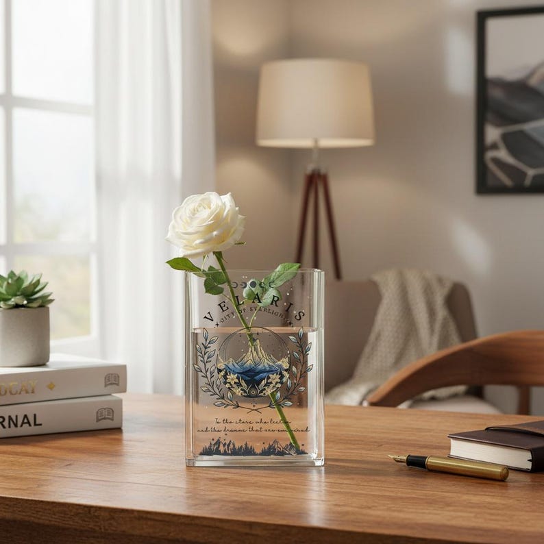Puede incluir: Un jarr&oacute;n de cristal transparente con una rosa blanca. El jarr&oacute;n rectangular presenta el texto "VELARIS CITY OF STARLIGHT" con un dise&ntilde;o de monta&ntilde;a y flores. El jarr&oacute;n est&aacute; sobre una mesa de madera.