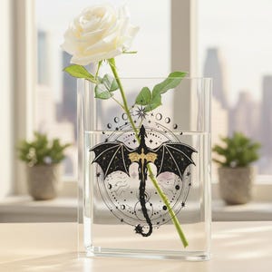 Puede incluir: Un jarrón rectangular transparente con un diseño de dragón negro y dorado. Una rosa blanca con hojas verdes se coloca dentro del jarrón. El diseño del dragón incluye un fondo celestial con estrellas y lunas. El jarrón está sobre una superficie clara.
