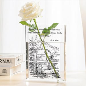 May include: A clear, rectangular glass vase holding a white rose. The vase has a black and white illustration of a boy and a bear, with a quote by A.A. Milne. The vase is filled with water. The rose stem is green.