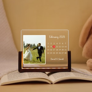 May include: A personalized wedding photo night light with a wooden base. The clear acrylic panel displays a photo of a couple, a calendar for February 2026, and the names "David & Sarah."