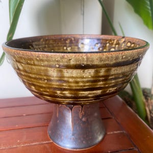 May include: A handcrafted ceramic bowl with a pedestal base. The bowl features a textured, earthy brown glaze with darker brown accents and a speckled pattern. The pedestal is a darker shade of brown.