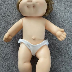 以下が含まれることがあります： 柔らかいビニール肌、青い目、薄茶色の髪のヴィンテージ人形。人形は白いおむつを履いています。人形の両腕は広げられており、笑顔です。