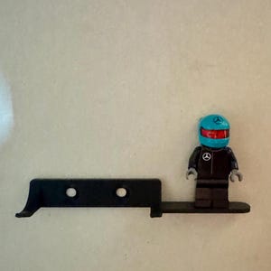 May include: A black Lego-style figure in a racing suit and helmet, standing next to a black, angular piece with two holes. The helmet is blue with a red visor and a logo. The figure is on a black base.