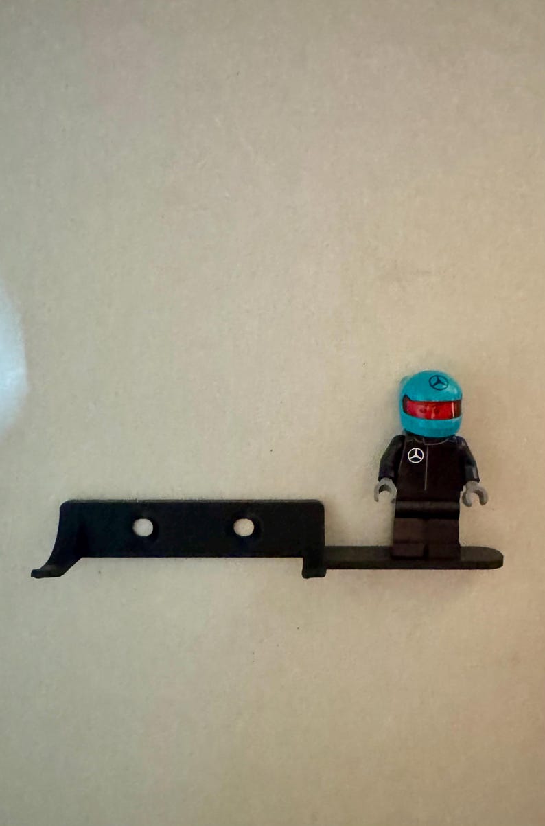 May include: A black Lego-style figure in a racing suit and helmet, standing next to a black, angular piece with two holes. The helmet is blue with a red visor and a logo. The figure is on a black base.