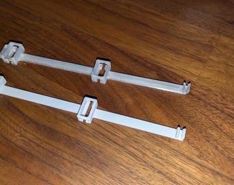 May include: Two light grey plastic straps, each featuring a rectangular sliding buckle and a small hook at one end. The straps have a textured surface and a square-shaped attachment point at the opposite end, designed for securing or fastening items.