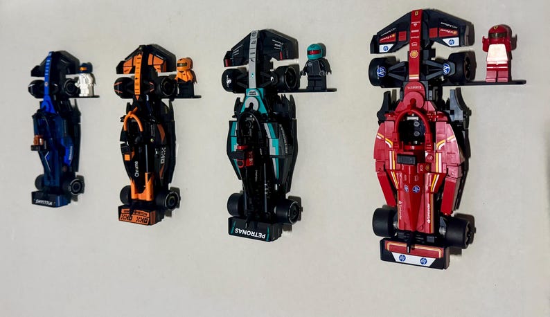 May include: Four toy Formula 1 race cars in blue, orange, teal, and red, mounted on a white wall. Each car has a unique design and is accompanied by a small figurine. The cars are displayed on small black shelves.