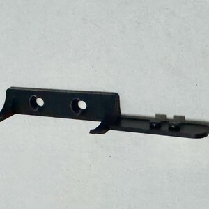 May include: A black plastic component with two circular holes and two curved extensions. The component has a rectangular shape with a series of small, raised rectangular features on one side. The object is set against a plain, light background.