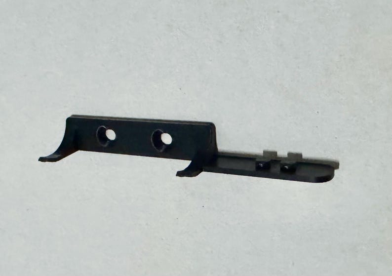 May include: A black plastic component with two circular holes and two curved extensions. The component has a rectangular shape with a series of small, raised rectangular features on one side. The object is set against a plain, light background.