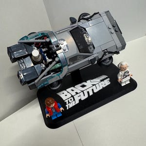 May include: A silver Lego DeLorean time machine model with detailed engine components, displayed on a black base with "Back to the Future" text. Includes Lego figures of Marty McFly and Doc Brown.