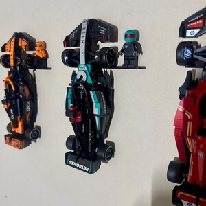May include: Three toy race cars, each mounted on a white wall. The central car is black and teal with the word "PETRONAS" visible. A small figure with a helmet stands next to it. The other cars are orange and red.