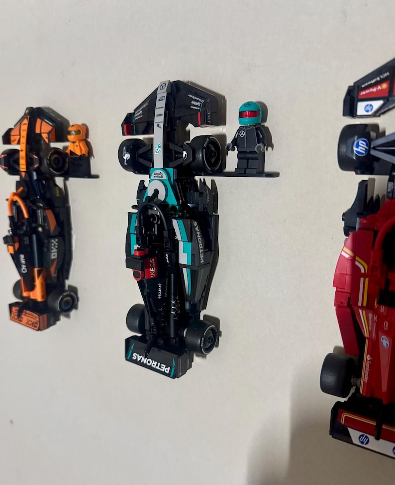 May include: Three toy race cars, each mounted on a white wall. The central car is black and teal with the word "PETRONAS" visible. A small figure with a helmet stands next to it. The other cars are orange and red.
