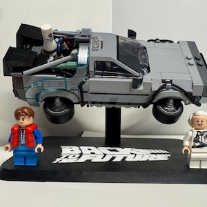 May include: A gray Lego DeLorean time machine model, with two Lego figures of Marty McFly and Doc Brown. The model is suspended above a black base with the words "Back to the Future" in white.