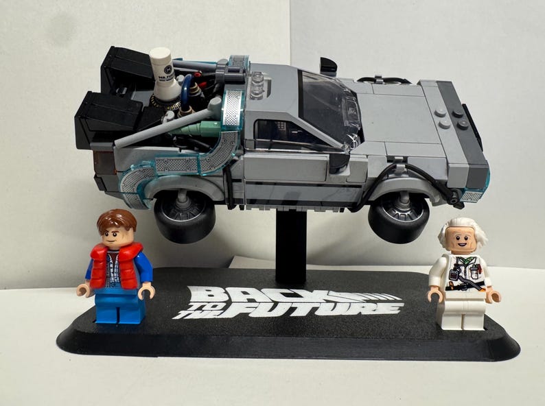 May include: A gray Lego DeLorean time machine model, with two Lego figures of Marty McFly and Doc Brown. The model is suspended above a black base with the words "Back to the Future" in white.