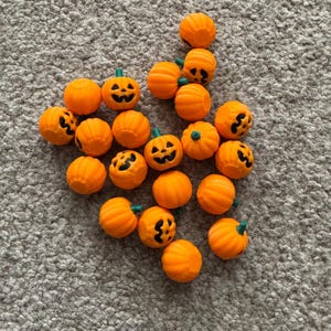 May include: A scattering of small, orange pumpkin decorations, each with a carved jack-o'-lantern face and a green stem. These Halloween-themed pumpkins are arranged on a textured, grey surface, ideal for seasonal crafts and decorations.