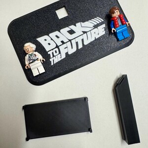 May include: A black display stand with the text "BACK TO THE FUTURE" in white. Two Lego figures of characters from the movie are on the stand. There are also two black rectangular pieces.