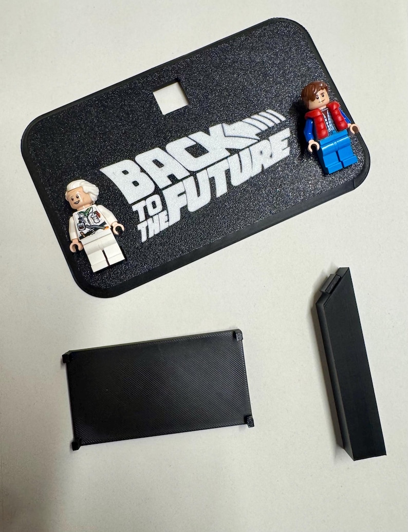May include: A black display stand with the text "BACK TO THE FUTURE" in white. Two Lego figures of characters from the movie are on the stand. There are also two black rectangular pieces.