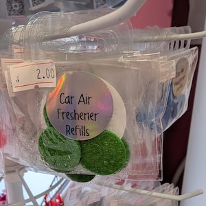 May include: Clear plastic packaging containing car air freshener refills. The refills are round, with green and white felt discs. The packaging is hanging on a white metal rack. The label reads "Car Air Freshener Refills".