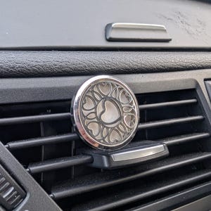 Könnte beinhalten: Ein silberner Auto-Luftauslassdiffusor mit herzförmigem Design. Der runde Diffusor hat ein dekoratives Muster aus ineinandergreifenden Herzen. Er ist an einem schwarzen Lüftungsschlitz befestigt.