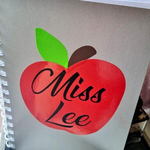 May include: A spiral-bound notebook with a gray cover featuring a large red apple graphic. The apple has a green leaf and a brown stem. The words "Miss Lee" are written in black script across the apple.