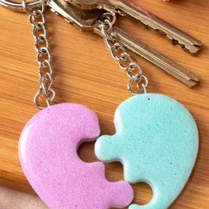 Puede incluir: Dos llaveros con piezas de rompecabezas en forma de corazón, uno lila claro, otro azul claro, ambos con purpurina. Están unidos a una cadena plateada y a un llavero con dos llaves.