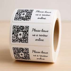 May include: A roll of white stickers featuring black QR codes and the text "Please leave us a review online." The stickers are rectangular with rounded corners. The roll is positioned on a light brown surface.