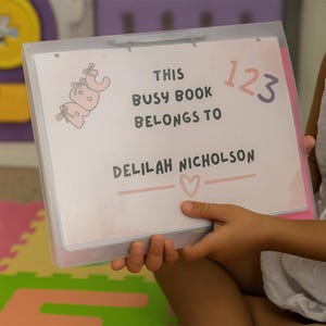 May include: A clear plastic binder holds pages with text and illustrations. The text reads "THIS BUSY BOOK BELONGS TO DELILAH NICHOLSON" with a heart symbol. A drawing of a pink elephant is in the upper left corner, and the numbers "123" are in the upper right.