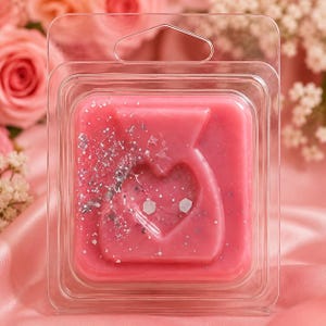 May include: A square pink wax melt in a clear plastic clamshell container. The wax features a molded heart shape in the center and is decorated with silver glitter particles scattered across the surface, including two small white hexagonal accents.