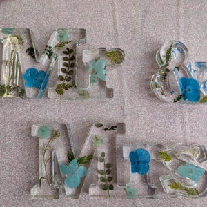 May include: Clear resin letters spelling "Mrs & Mrs", each embedded with dried flowers in shades of blue, green, and gold. The letters are displayed on a light pink, glittery surface.