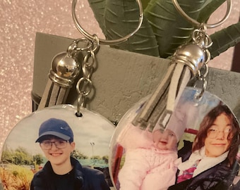 May include: Two round photo keychains with silver key rings and tassels. One keychain features a photo of a young person wearing a blue hat and glasses. The other keychain features a photo of a young child in a pink outfit being held by a person with long brown hair.