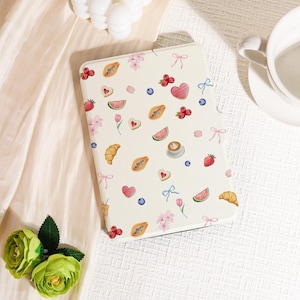 Può includere: Una custodia per tablet color crema ornata da un motivo di illustrazioni ad acquerello. Il design include croissant, cuori, bacche, fette di papaya e fiocchi. La custodia è rettangolare e ha angoli arrotondati.