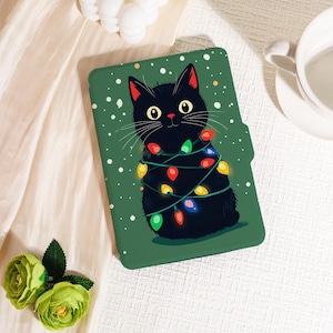 May include: Green tablet case with a black cat design wrapped in colourful Christmas lights. The cat has yellow eyes and red inner ears. White dots are scattered on the green background, creating a festive holiday theme.