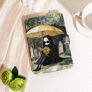 May include: A tablet case featuring a watercolor illustration of a woman in black attire and sunglasses, reading a book under a yellow umbrella in a graveyard. The book reads "How to Add People".