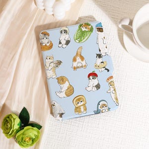 May include: A light blue tablet case decorated with cartoon cat illustrations. The cats are shown in various poses and outfits, such as a chef's hat, a shark costume, and a red beret. The case is set against a white backdrop, with a green floral arrangement.