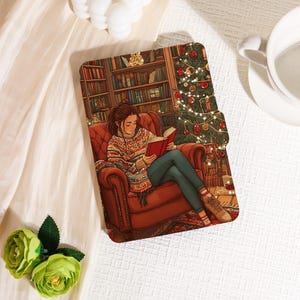 May include: A decorative tablet case with an illustration of a person reading a book in a cosy room with a Christmas tree and bookshelves. The person is wearing a colourful sweater and jeans, sitting in a red armchair.