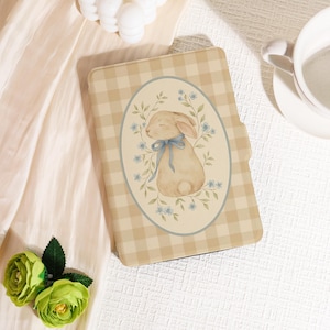 May include: A beige tablet case with a gingham pattern. The case features an oval design with a watercolor illustration of a bunny wearing a blue bow, surrounded by blue flowers and green leaves. The case is closed.