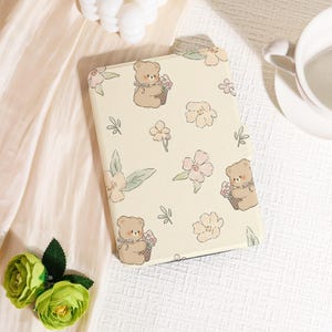 May include: A cream tablet case with a repeating pattern of cartoon teddy bears holding flower baskets and delicate pink and white floral designs. The case is rectangular and has a smooth surface.