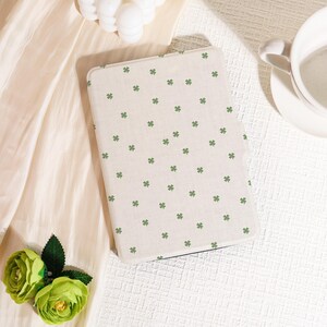 May include: A cream-coloured tablet case with a pattern of small green shamrocks. The case is rectangular with rounded corners. The background includes a white teacup, folded fabric, and green floral accents.