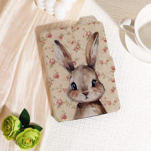 Può includere: Una custodia beige per tablet con un'illustrazione ad acquerello di un coniglio. La custodia ha un motivo floreale con rose rosse e rosa. Il coniglio ha orecchie lunghe e grandi occhi. La custodia è chiusa.