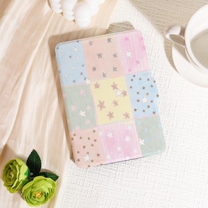 May include: A pastel-colored tablet case with a patchwork design. The case features squares of light blue, pink, yellow, and green, each adorned with star patterns. A cup of coffee and a small bouquet of green flowers are in the background.