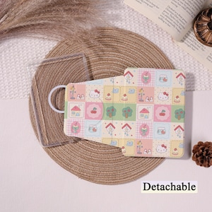 May include: A detachable tablet case with a colourful patchwork design featuring houses, trees, cherries, and Hello Kitty characters. The case is light-coloured with a clear plastic frame.