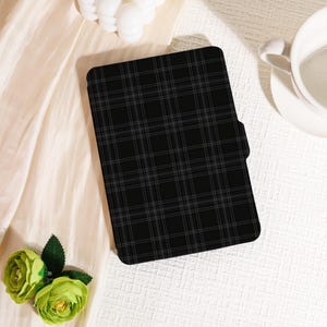 May include: A black tablet case with a gray plaid pattern. The case is rectangular with rounded corners and a side pocket. The background includes a white teacup, a light-colored fabric, and green flowers.