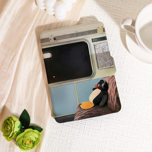 Puede incluir: Una funda de teléfono con un pingüino de dibujos animados con auriculares, sentado en un vagón de tren. La funda tiene un esquema de color azul claro y beige, con un detalle de mapa. El pingüino tiene una nariz roja y pies amarillos.